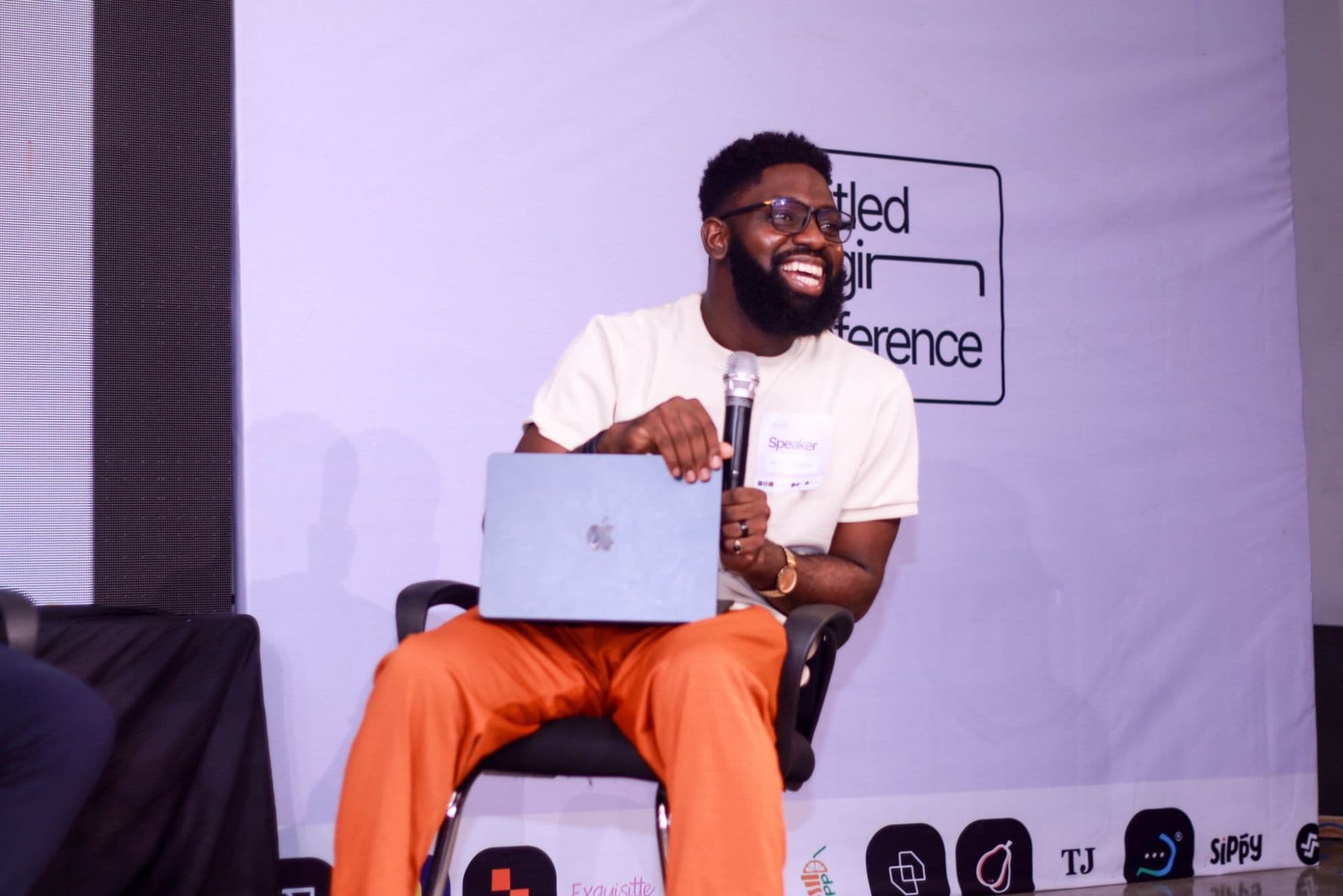 Alex on stage as a conference speaker, laughing, with a microphone and laptop